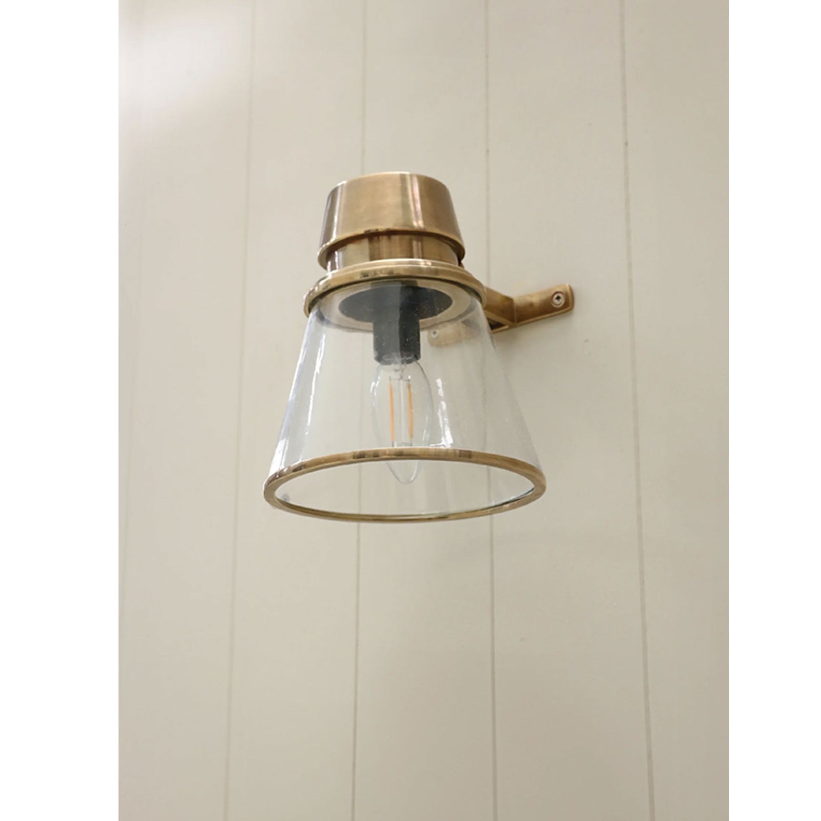 Wall-mounted light fixture with brass finish and clear glass shade on a white paneled wall.