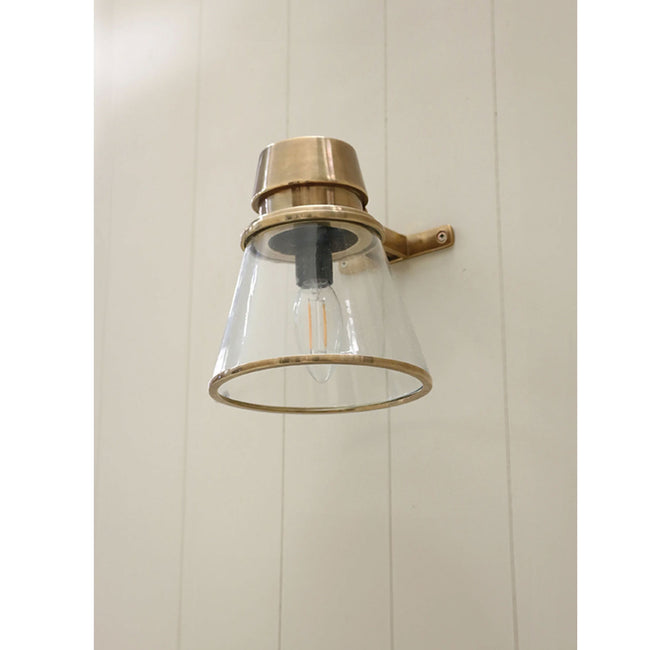 Wall-mounted light fixture with brass finish and clear glass shade on a white paneled wall.